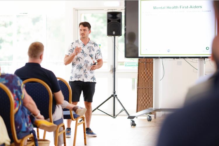 Mark Wheeler speaking to a seated room, a screen behind him titled Mental Health First-Aiders
